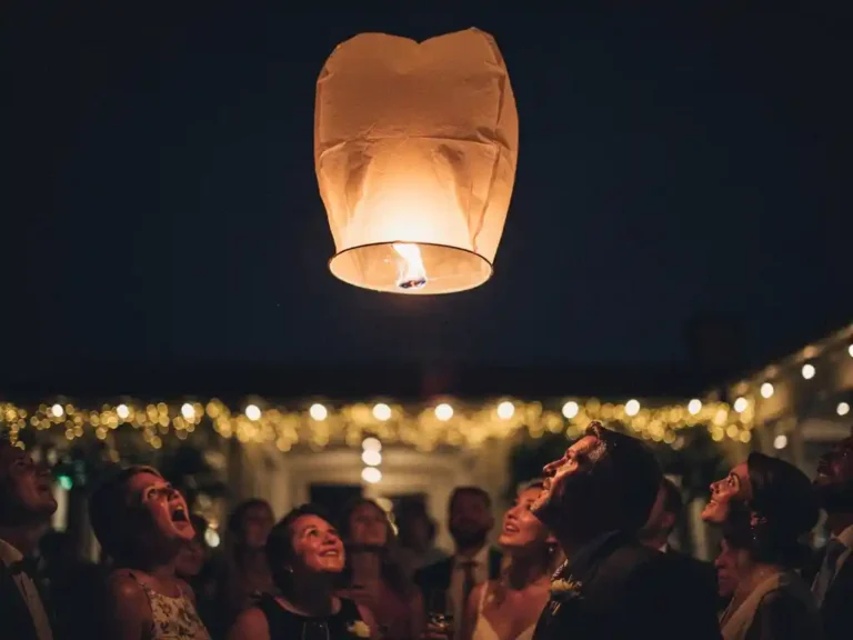 Oplichtende lantaarn stijgt op in donkere avondlucht boven silhouetten van gasten bij een buitenevenement, warme gouden gloed op omhooggerichte gezichten.